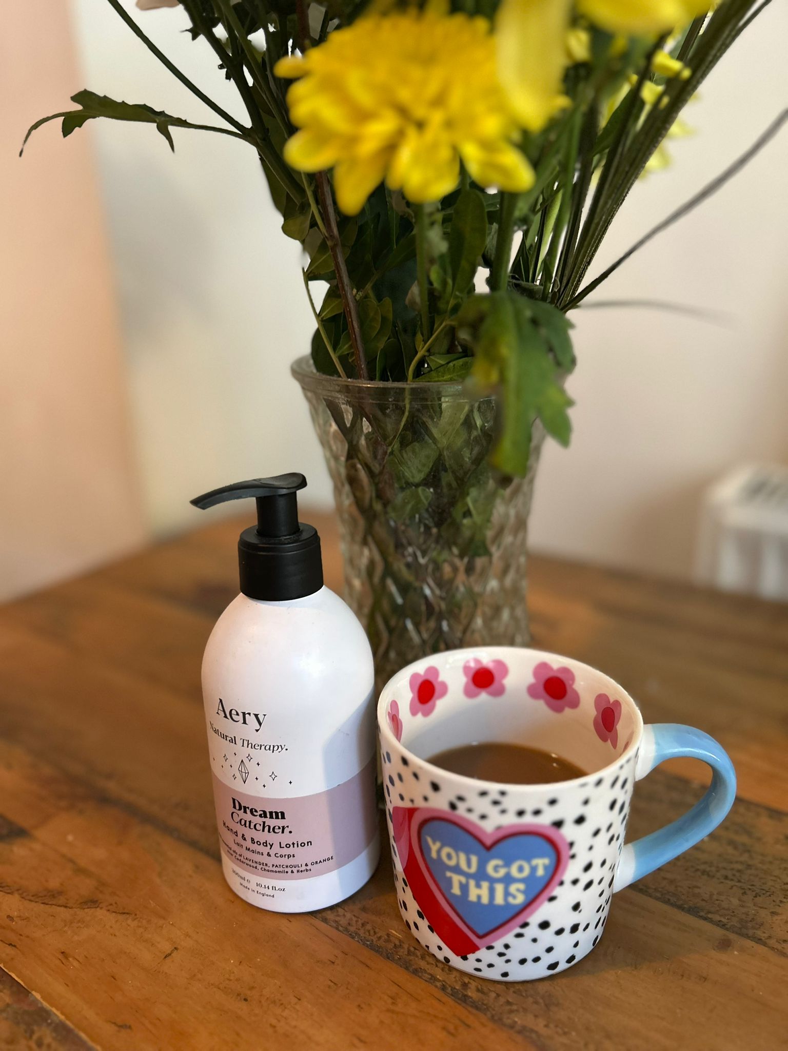 Lotion bottle, mug with 'You Got This' design, and vase with yellow flowers on a wooden surface.